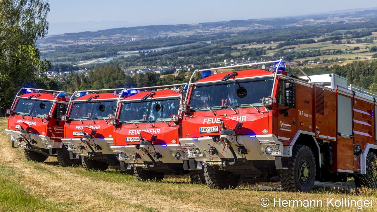 Feuerwehr-Löschfahrzeuge in Reih und Glied vor einer Landschaft mit Fluss und Ort